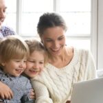 Smiling family of mom, dad, and two children on a couch watching digital video on a laptop in a bright room in front of the window