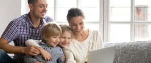 Smiling family of mom, dad, and two children on a couch watching digital video on a laptop in a bright room in front of the window