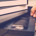 Someone pushing a VHS tape into a VHS player, with more tapes on top of the player.