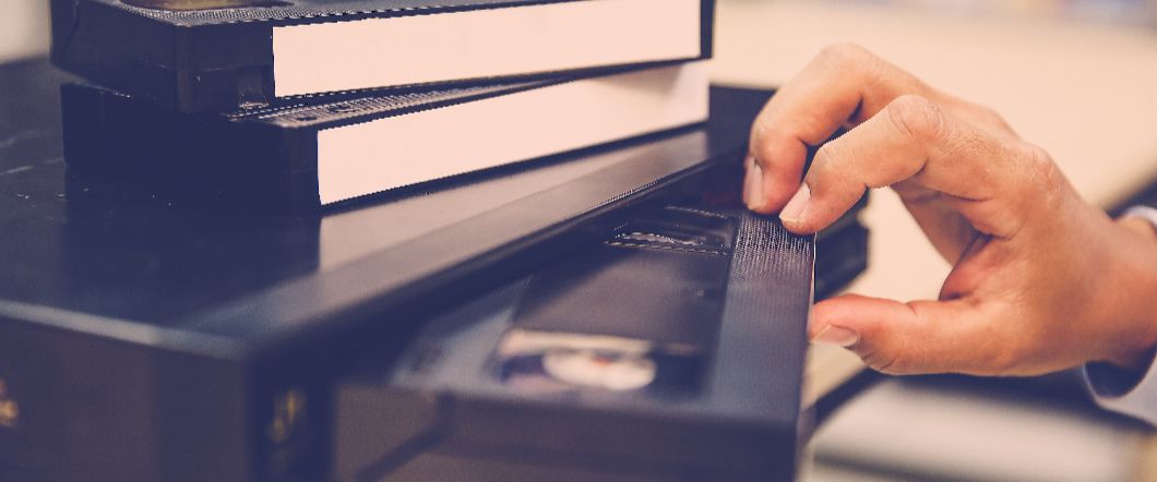 Someone pushing a VHS tape into a VHS player, with more tapes on top of the player.